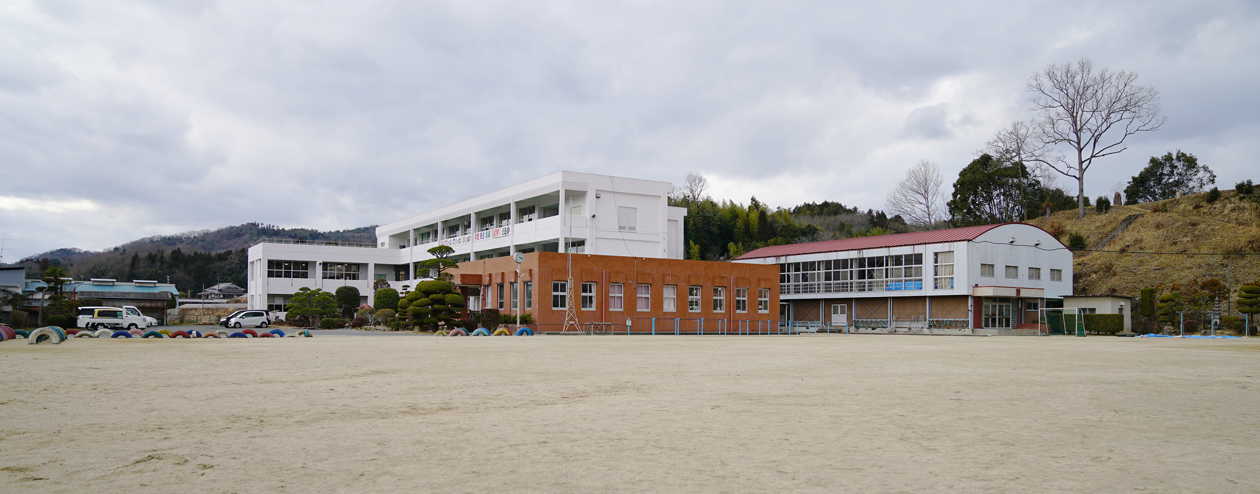 カラフルな色のタイヤとびや鉄棒などの遊具が設置されている運動場から、2階建てと3階建ての校舎や、赤色の屋根をした体育館を撮影した和気町立佐伯小学校の写真