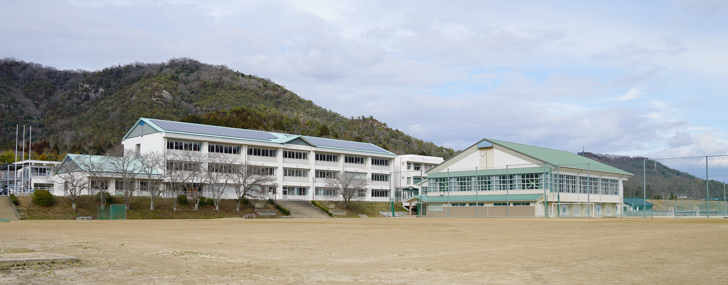 山の麓に建つ3階建ての校舎と緑色の屋根の体育館を運動場から撮影した和気町立和気中学校の写真