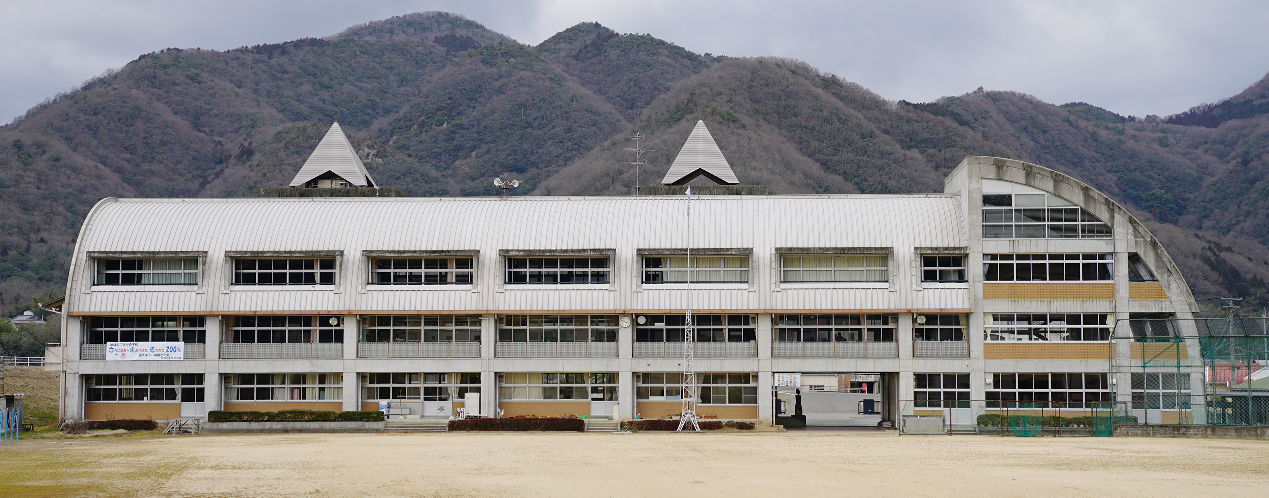 横に広がる山並みの麓に建つ、屋上の左右に三角錐の様な形をした物が設置された3階建ての佐伯中学校の校舎を運動場から撮影した写真