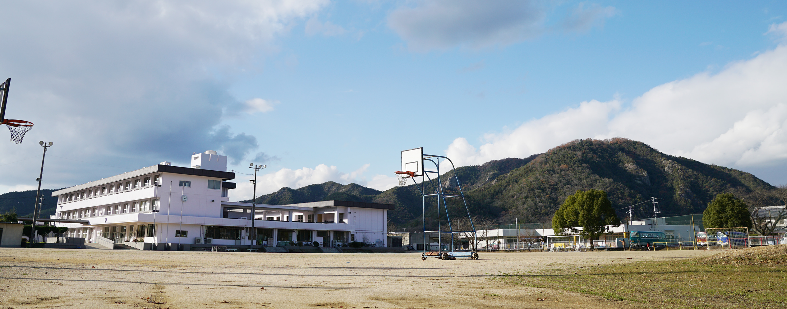 3階建てと2階建ての校舎が渡り廊下で繋がっており、可動式のバスケットゴールが設置されている運動場から撮影した和気町立和気小学校の写真