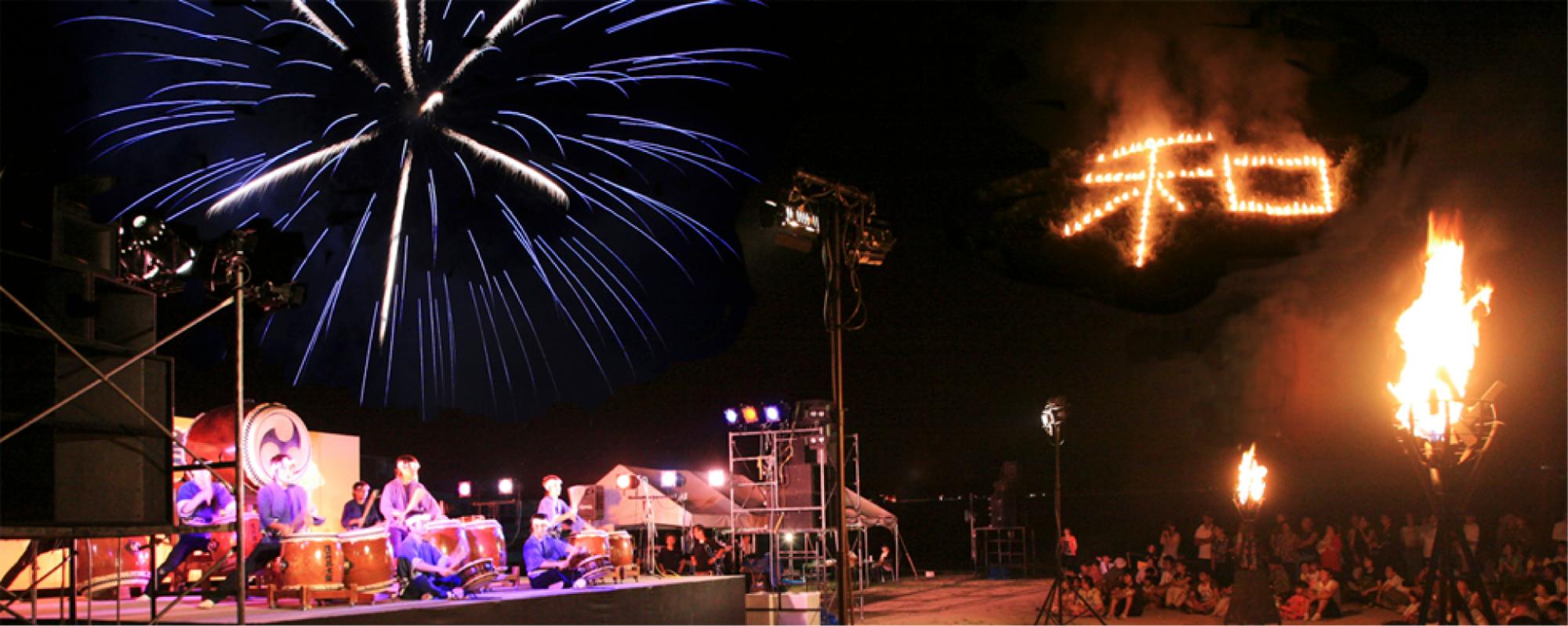 高い位置に「和」の火文字、空には大きな青い花火が浮かびあがっており、地上の舞台で法被を着た人達が太鼓を演奏を行っている様子を大勢の参加者が見ているお祭りの様子の写真
