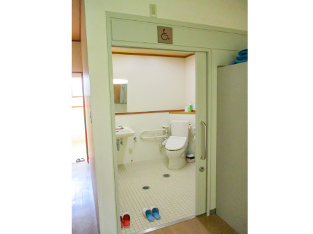 トイレのドアが空いていて、赤と青のスリッパが並んで置かれ、右奥にバリアフリー付きの便器がありその左には鏡と手洗い場が見えるトイレの写真