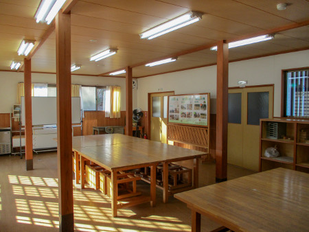 木造の支柱や長方形のテーブルとその下に収納された椅子、陽に照らされた窓の格子状の影が綺麗に床にうつっている室内の写真
