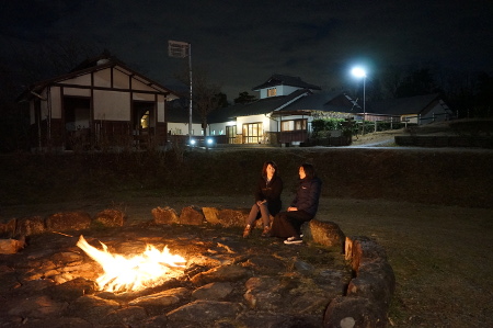 遠くでうっすらと外灯がついている薄暗い中、円形に並べられた大きな石の中心で木が燃えており、その近くで女性2人が石に座って話しをしている写真