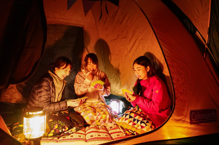 夜のテントにランタンを点け女性3人がトランプしている写真
