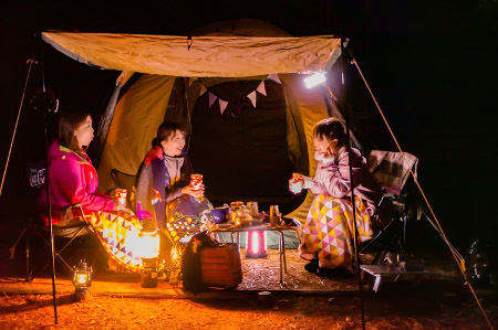 暗闇の中、テントの外のタープの下でランタンを点け、テーブルには飲み物、食べ物が置かれ女性3人が談笑している写真