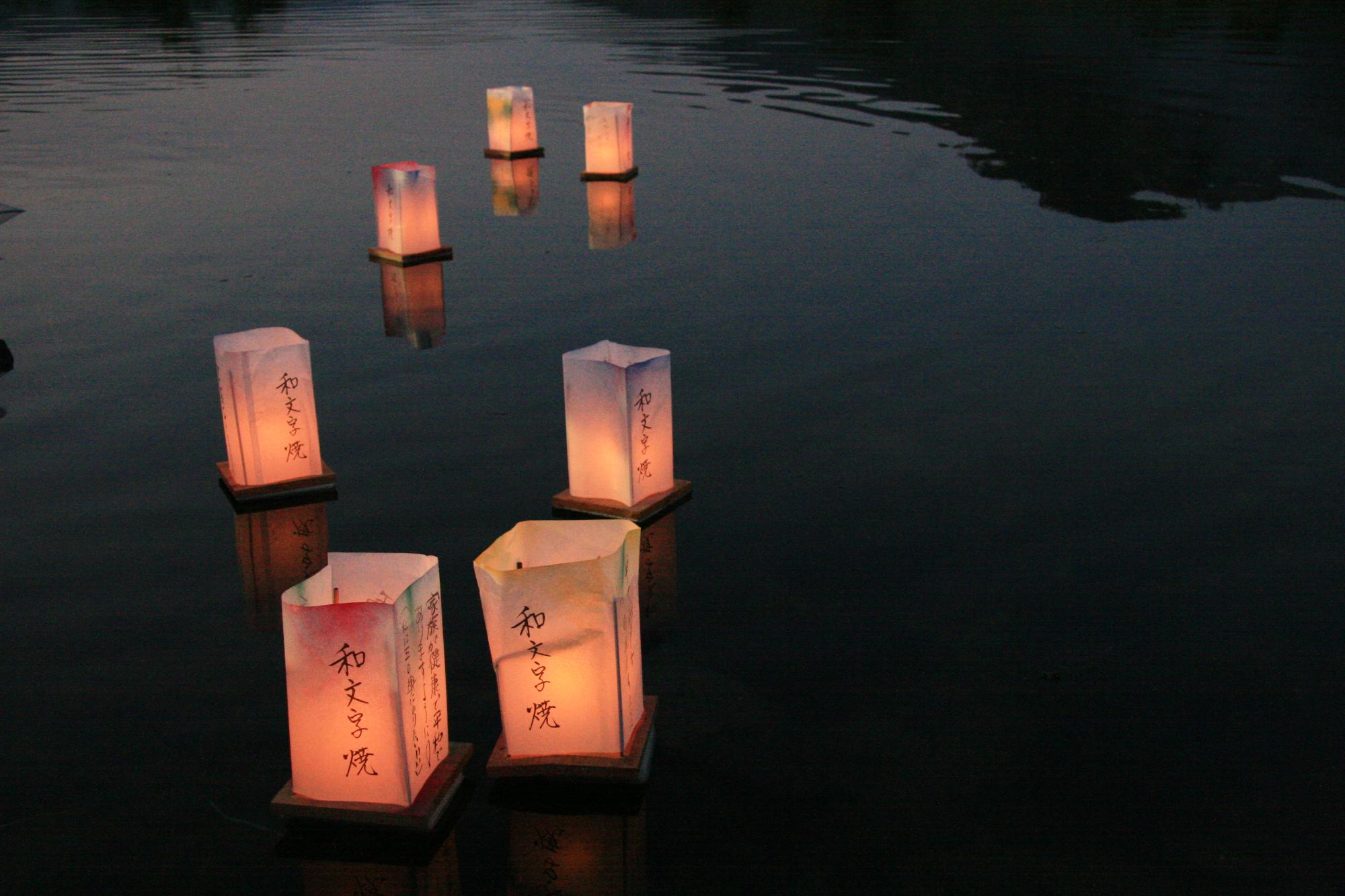 「和文字焼」の文字が書かれた明かりのついた灯ろうが7つ水の上に浮かんでいる写真