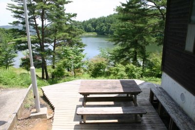 池を望むコテージの外のウッドデッキにバーベキューが楽しめるテーブルや椅子が設置されている写真
