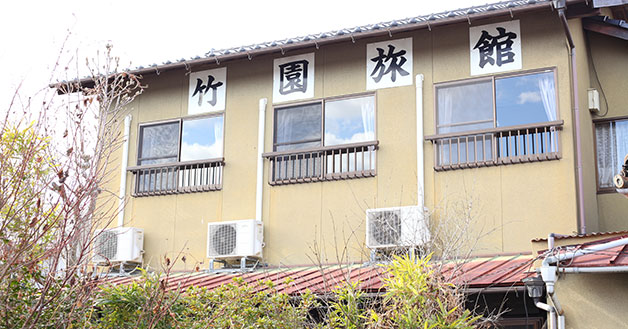 屋根の下に一文字ずつ「竹園旅館」と表示された、ベージュ色の外壁のビジネス旅館竹園の建物外観の写真
