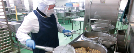 白色の作業着に紺色のエプロンを着用したスタッフが、大きな鍋で調理を行っている写真