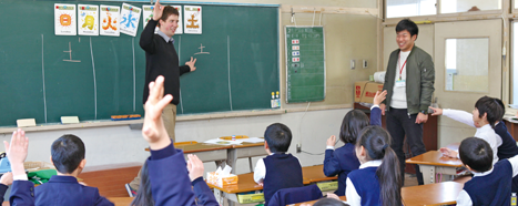 黒板に曜日が書かれた用紙が貼られ、黒板の前にALTの男性教師が立ち、自席に座った児童たちと右手を挙げた同じポーズをとっている授業の様子の写真