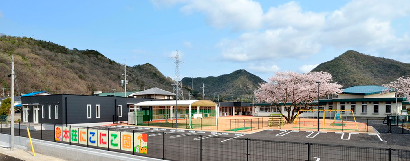 小高い山の麓の敷地を囲むフェンスに一文字ずつカラフルな色で「本荘にこにこ園」と書かれ、敷地内に駐車場、ブランコなどの遊具が設置され満開の花が咲く2本の桜の木が立つ園庭、奥に園舎が建っている写真