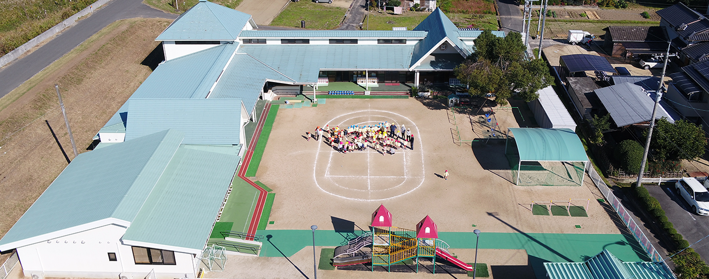 フェンスで囲まれた敷地内左側と奥にL字型の薄緑色の屋根の園舎が建ち、右側に一本の大きな木や鉄棒、手前にピンク色の屋根をした複合型遊具が設置され、中央の白線がひかれた運動場に先生や園児たちが集まっている佐伯にこにこ園を上空から撮影した写真