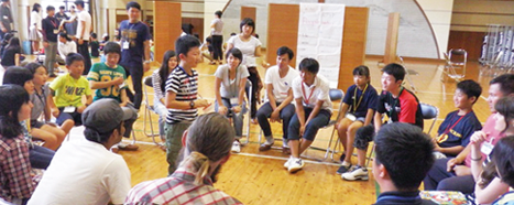 室内の広場で、円形に設置されたパイプ椅子に参加者が座り、中央に男の子が立っているイングリッシュキャンプのゲームの様子の写真