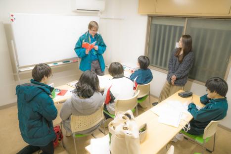 ホワイトボードの前に教科書を持った外国人講師が立ち、子供が前列に3名、後列に1名座った室内で授業を行っている様子の写真